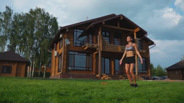 Slanke mooie vrouw in sportkleding op de achtergrond van het huis op het gazon voert sprongen voor cardio-training en vetverbranding - Video
