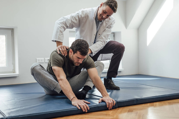 Young handsome Caucasian man stretching his back with his physician helping. - Photo, image