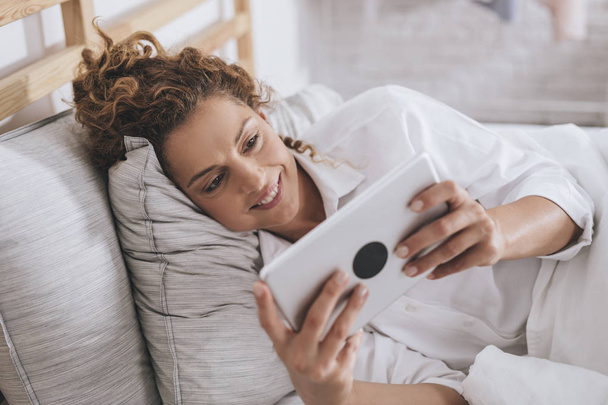 Pretty smiling Caucasian woman lying in her bed and reading on a tablet. - Foto, Imagem