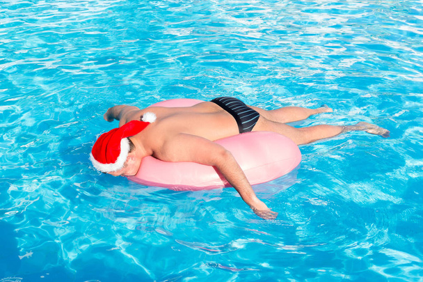 ένας μεθυσμένος άντρας στην Αϊ Βασίλη καπέλο κολυμπά σε φουσκωτά κύκλος στην πισίνα. ταξίδια από μια ρωσική τουριστική. - Φωτογραφία, εικόνα