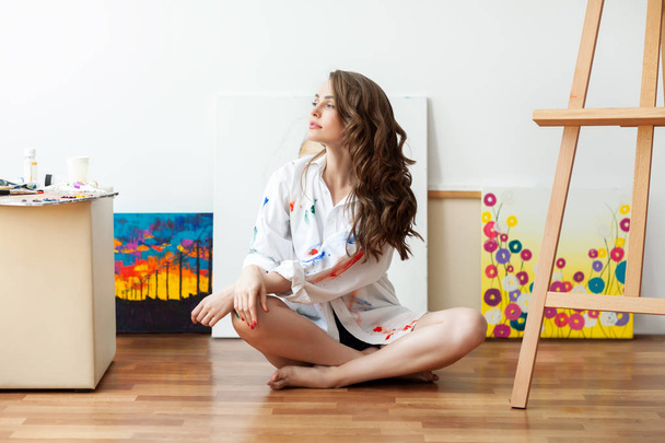 Pretty woman painter sitting on floor in front of canvas and drawing in art studio - Foto, Imagen