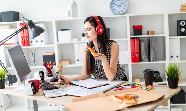 Ein junges Mädchen steht neben einem Tisch, hält einen grünen Filzstift und ein Stück Pizza in der Hand. vor dem Mädchen auf dem Tisch liegt eine Magnettafel. auf dem Kopf des Mädchens mit Kopfhörer. - Foto, Bild
