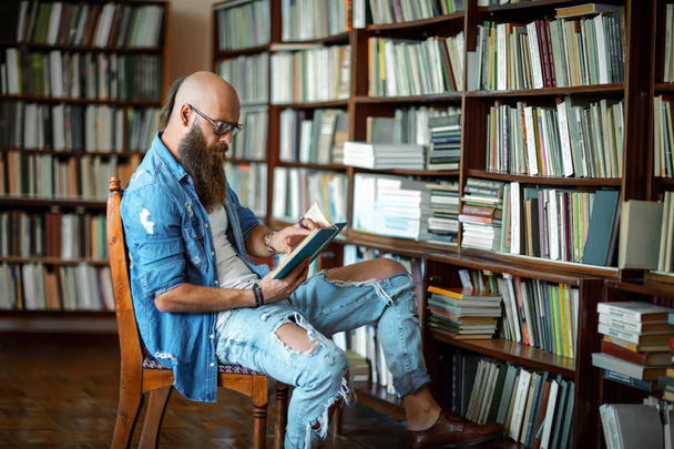 Hipster γενειοφόρος φοιτητής που σπουδάζει στη βιβλιοθήκη στο Πανεπιστήμιο που κάθονται σε παλιά καρέκλα - Φωτογραφία, εικόνα