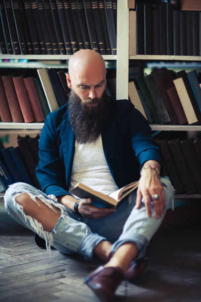 Handsome bearded man sitting on floor, reading book and relaxing in library or bookstore - Фото, зображення