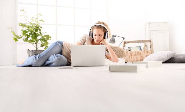 junge Frau mit Computer, Kopfhörer, Smartphone und Büchern, auf dem Boden liegend im Wohnzimmer vor weißem Fenster im Hintergrund - Foto, Bild