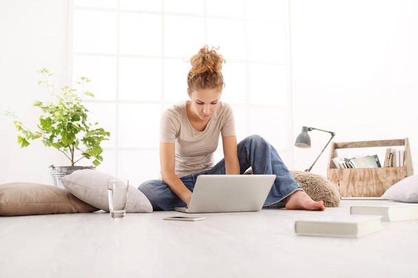 junge Frau mit Computer, Smartphone und Büchern, auf dem Boden im Wohnzimmer sitzend, im Hintergrund ein weißes Fenster - Foto, Bild