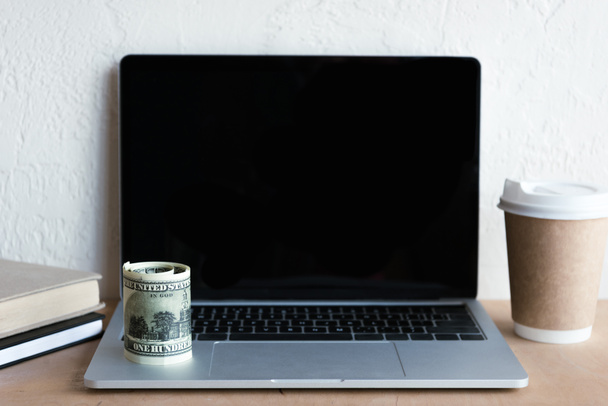 close-up view of laptop with blank screen, rolled dollar banknotes, books and coffee to go on table - 写真・画像
