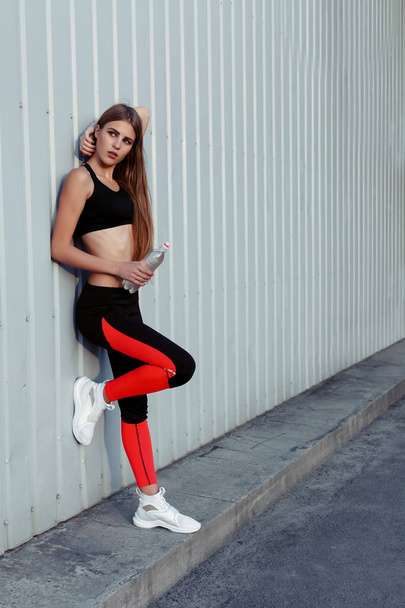 Female athlete drinking water while standing by a grey wall. Slim woman in sportswear taking a break after workout. - Photo, image