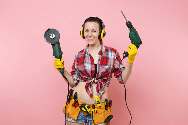 手袋でエネルギーの便利屋女性, ノイズ絶縁ヘッドフォン、保持力の楽器の完全キット ツール ベルト見たピンクの背景に分離された電気ドリル。男性の仕事の女性。改修工事コンセプト - 写真・画像