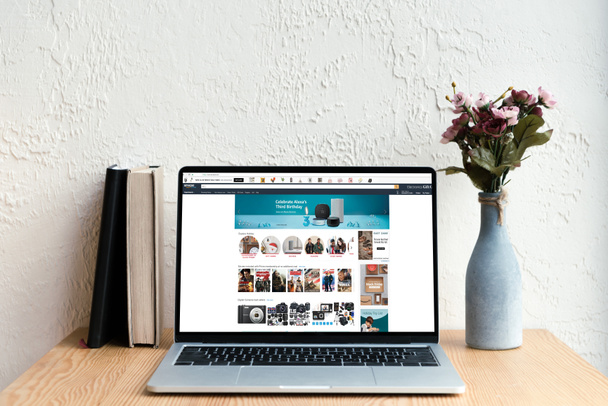 laptop with amazon website on screen, books and flowers in vase on wooden table - Valokuva, kuva