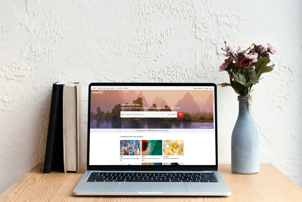 laptop with shutterstock website on screen, books and flowers in vase on wooden table - 写真・画像