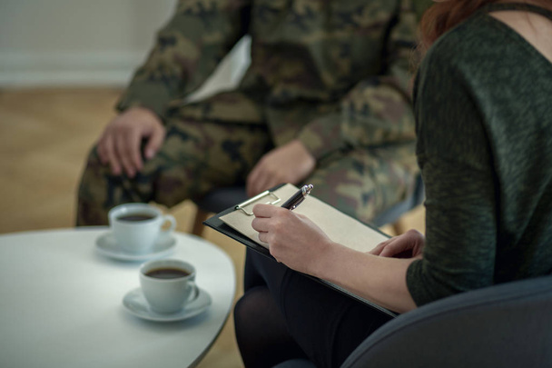 Close-up of psychologist writing down notes while talking to her patient from army - Фото, изображение