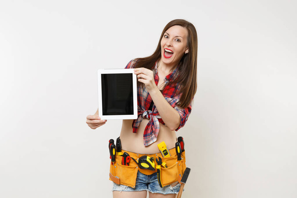 Frau im Bausatz Werkzeuge Gürtel voller Vielfalt Instrumente halten Tablet-PC mit leerem schwarzen Bildschirm Touchscreen auf weißem Hintergrund isoliert. Frauen, die männliche Arbeit verrichten. Sanierungskonzept - Foto, Bild