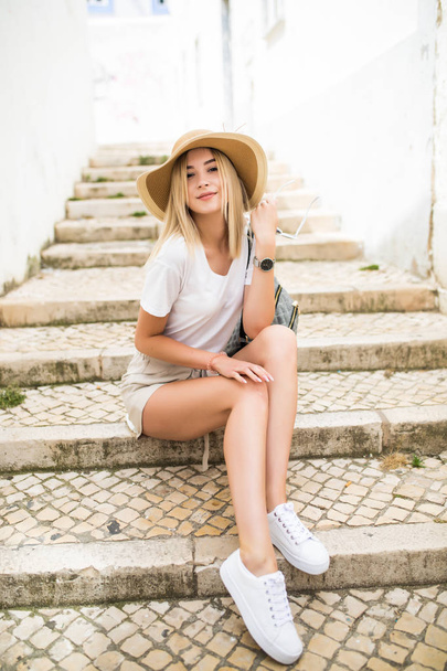 Ευτυχισμένη γυναίκα με καπέλο καλοκαίρι κάθομαι στις σκάλες σε δρόμο της πόλης - Φωτογραφία, εικόνα