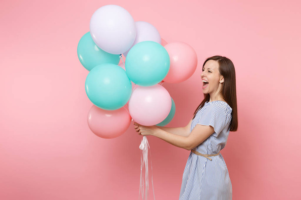 Portrait of joyful laughing young happy woman wearing blue dress holding colorful air balloons looking aside isolated on bright pink background. Birthday holiday party, people sincere emotion concept - Foto, imagen