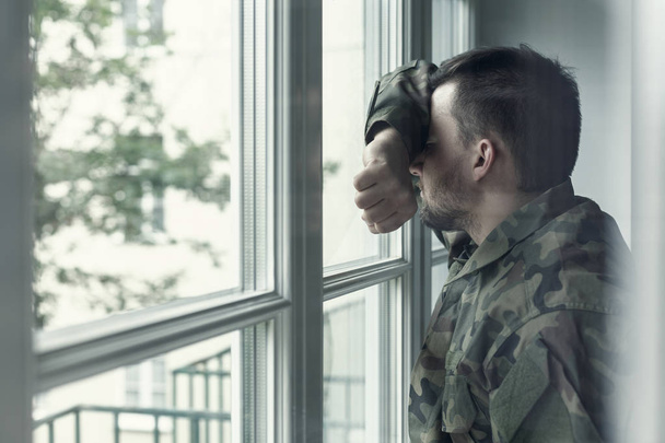 Soldado deprimido e triste em uniforme verde com trauma após a guerra em pé perto da janela
 - Foto, Imagem