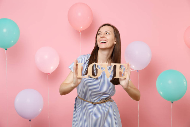 Portrait of laughing attractive young woman in blue dress holding wooden word letters love on pastel pink background with colorful air balloons. Birthday holiday party people sincere emotions concept - Photo, Image