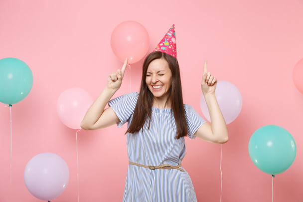 Portrét radost mladé ženy se zavřenýma očima v papírovou čepicí a modré šaty zvýrazňující ukazováček na růžovém pozadí s balónky barevné vzduchu. Dovolená oslava narozenin, upřímné emoce lidí - Fotografie, Obrázek