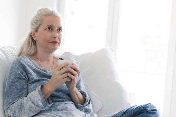 Senior woman sitting in livingroom with cup in her hands. - Fotoğraf, Görsel