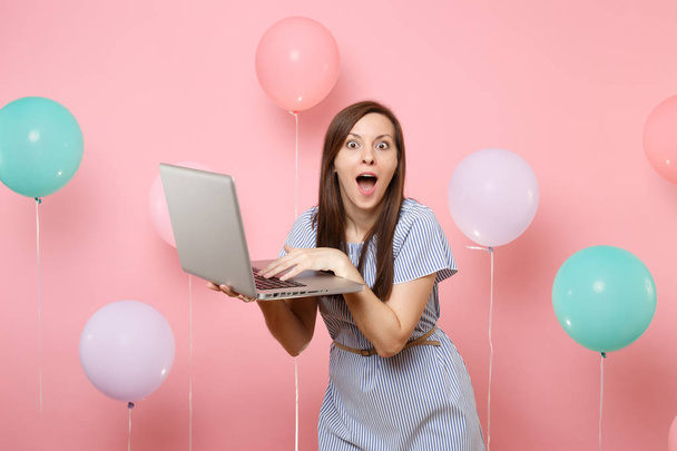 Portrait of shocked young woman with opened mouth in blue dress holding using laptop pc computer on pink background with colorful air balloons. Birthday holiday party people sincere emotions concept - Photo, Image