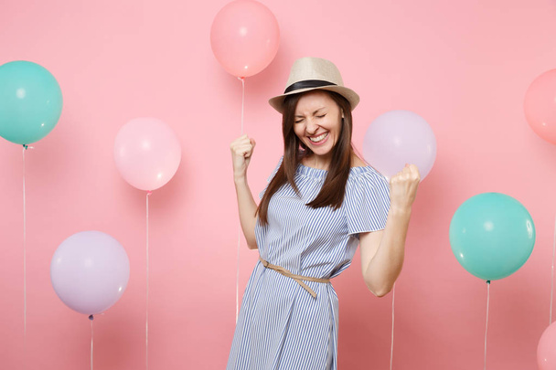 Portrait of joyful pretty young woman wearing straw summer hat and blue dress do winner gesture saying Yes on pink background with colorful air balloon. Birthday holiday party people sincere emotions - Fotó, kép