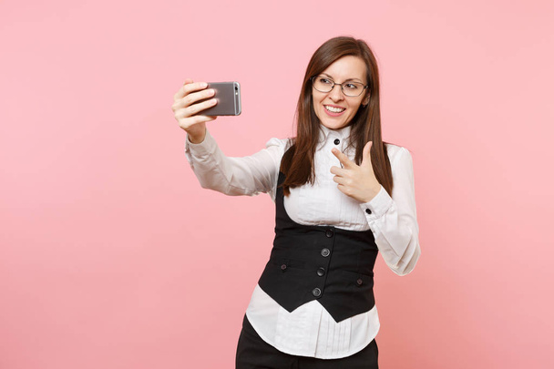 Молодая улыбающаяся деловая женщина в очках делает селфи-снимок на мобильном телефоне, указывая указательным пальцем на розовый фон. Леди-босс. Достижение карьерного богатства. Копирование места для рекламы
 - Фото, изображение