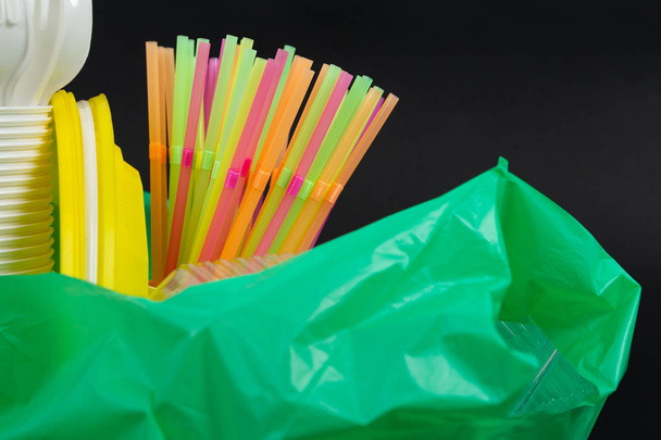 Closeup of colorful plastic waste in a green garbage bag as recyclable single use cutlery pollution junk concept isolated on black background - Foto, Imagen