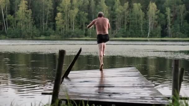 Взрослый кавказский мужчина прыгает с деревянного пирса в озеро
. - Кадры, видео