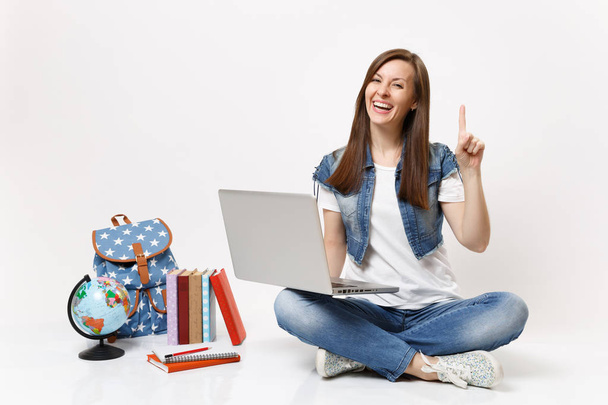 Nuori nauraa nainen opiskelija pitämällä kannettava tietokone tietokone osoittaa etusormella istuu lähellä maailmaa, reppu, koulukirjat eristetty valkoisella taustalla. Koulutus lukion yliopisto college
 - Valokuva, kuva