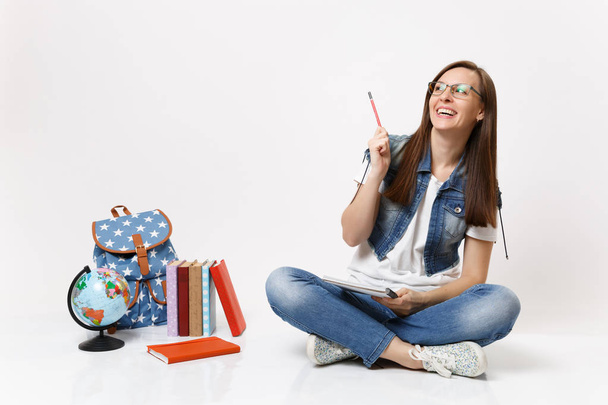 Młody szczęśliwy roześmiany student Kobieta, w okularach, wskazując ołówek się gospodarstwa notebook siedzi w pobliżu globe, plecak, podręczników szkolnych na białym tle na białym tle. Edukacja w liceum university college - Zdjęcie, obraz