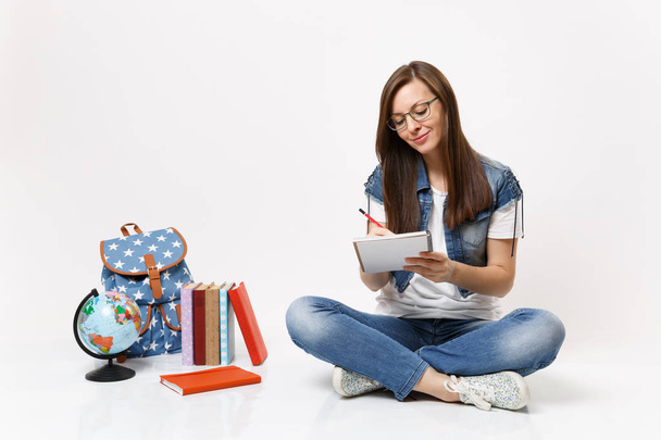 Молодая симпатичная улыбающаяся студентка в очках пишет заметки на блокноте, сидящем возле глобуса, рюкзаке, школьных книгах, изолированных на белом фоне. Образование в вузе
 - Фото, изображение