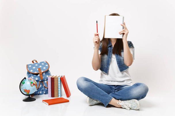 Young woman student in denim clothes holding pencil covering face with notebook sitting near globe, backpack, school books isolated on white background. Education in high school university college - Photo, Image