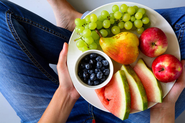 Woman's hands holding plate with summer fruits: watermelon, blueberry,apples,pear and grape on white plate. Healthy eating concept. Top view - Foto, immagini