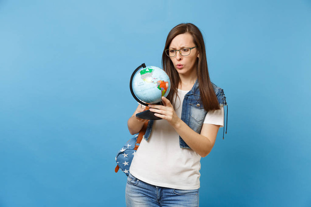 Портрет молодой озадаченной обеспокоенной студентки в очках с рюкзаком, смотрящей на мировой шар, ищущей страны, изолированные на синем фоне. Образование в вузе
 - Фото, изображение