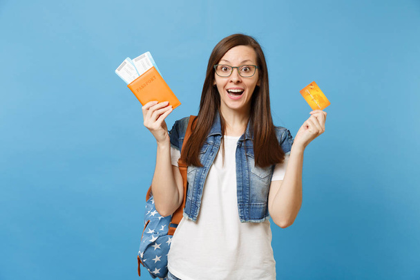 Young shocked excited woman student with backpack spread hands holding passport boarding pass ticket credit card isolated on blue background. Education in university college abroad. Air travel flight - Photo, Image