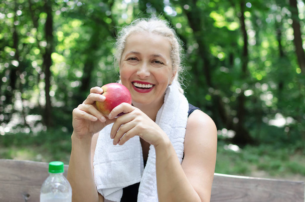 Πορτραίτο του χαμογελώντας μήλων εκμετάλλευσής ανώτερων γυναίκα, κάθεται στο πάρκο. Βλέπουν τα φωτογραφικών μηχανών. - Φωτογραφία, εικόνα