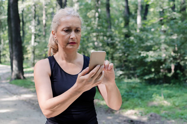 Portrait of senior woman holding and looking at phone in nature during work-out. - Фото, изображение