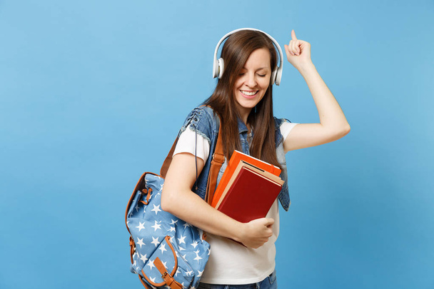 Fiatal aranyos nő diák farmer ruha, hátizsák fejhallgató figyelj zene tart iskolai könyv mutató mutatóujj tánc elszigetelt kék háttér. Középiskola Egyetem főiskolai oktatás - Fotó, kép