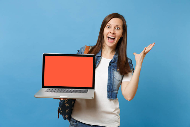 Young excited woman student with opened mouth spreading hands holding laptop pc computer with blank black empty screen isolated on blue background. Education in college. Copy space for advertisement - Photo, Image