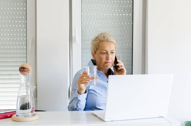 Senior beautiful woman working at home on laptop and talking on cellphone with glass in hand. - Photo, Image