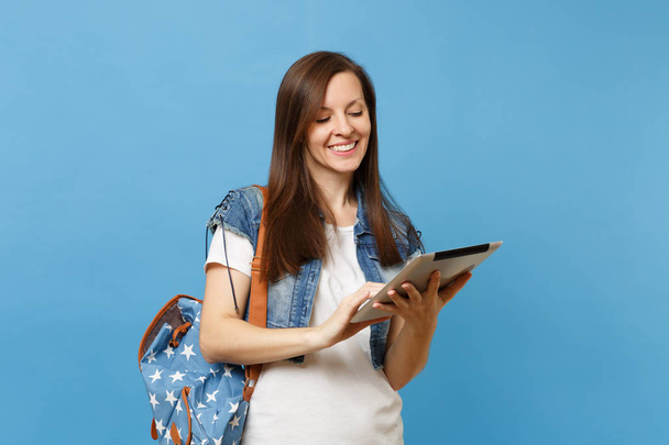Young pleasant beautiful woman student in denim clothes with backpack holding and using working on tablet pc computer isolated on blue background. Education in college. Copy space for advertisement - Photo, Image