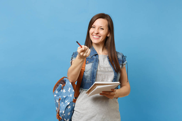 Portrait of young joyful smiling woman student in denim clothes with backpack hold notebook, pointing pencil on camera isolated on blue background. Education in high school university college concept - Photo, image