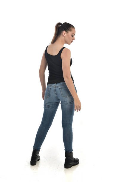 full length portrait of brunette girl wearing black single and jeans. standing pose, back to the camera. isolated on white studio background. - Foto, Imagen