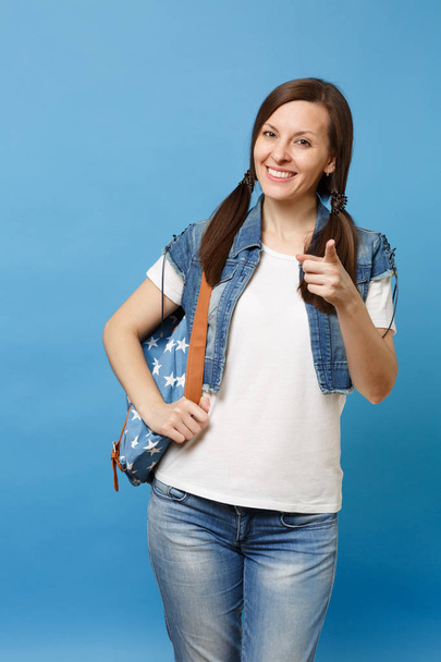 Portrait of young pleasant lovely woman student in denim clothes with backpack pointing index finger on camera isolated on blue background. Education in high school. Copy space for advertisement - Fotoğraf, Görsel