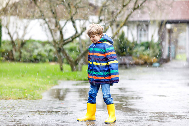 Little kid boy wearing yellow rain boots and walking during sleet, rain and snow on cold day. Child in colorful fashion casual clothes jumping in a puddle. Having fun outdoors. - Photo, Image