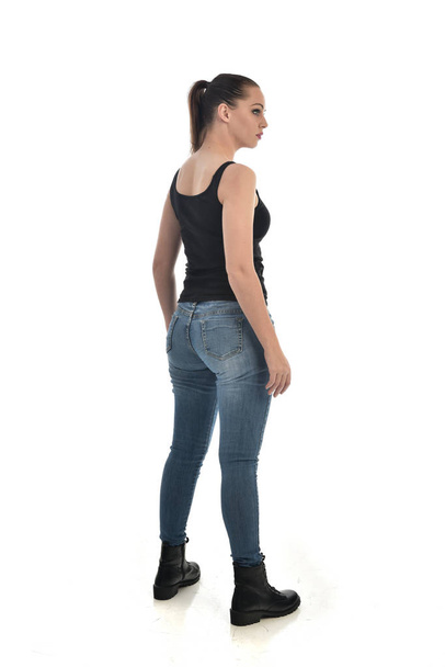 full length portrait of brunette girl wearing black single and jeans. standing pose, back to the camera. isolated on white studio background. - Zdjęcie, obraz