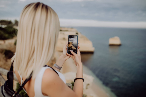Young beauty woman take photo on the phone of beautiful ocean landscape with water and cliff. - Photo, Image