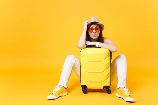 Bored tourist woman in summer casual clothes hat sit with suitcase, wait for plane isolated on yellow orange background. Girl traveling abroad to travel on weekends getaway. Air flight concept - Fotografie, Obrázek