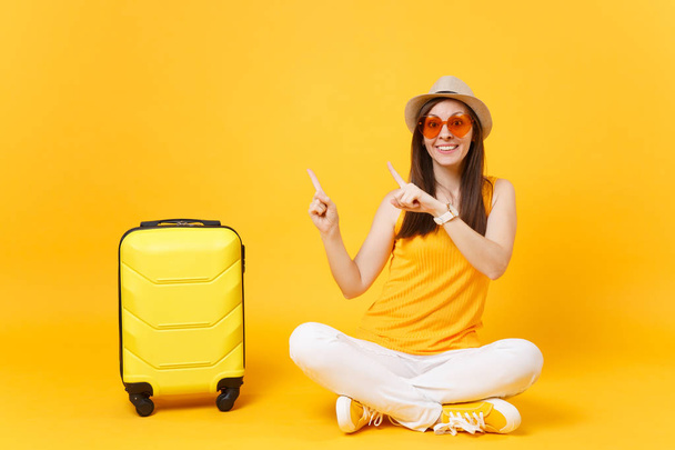 Traveler tourist woman in summer casual clothes, hat sit with suitcase isolated on yellow orange background. Female passenger traveling abroad to travel on weekends getaway. Air flight concept - Photo, Image