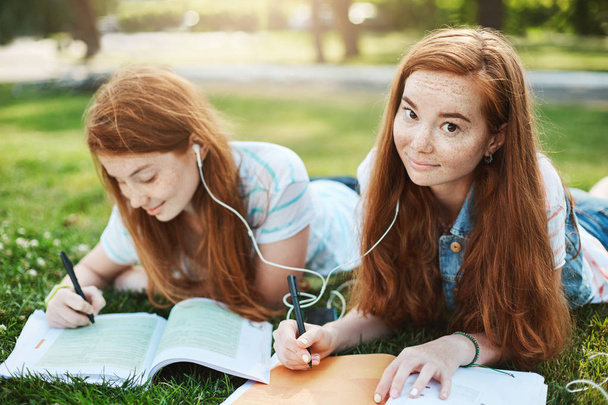 Charming redhead female with ginger hair gazing at camera with lifted eyebrows and cute smile, lying on grass in city park with sister, sharing earphones to listen music together and doing homework - Foto, Imagen